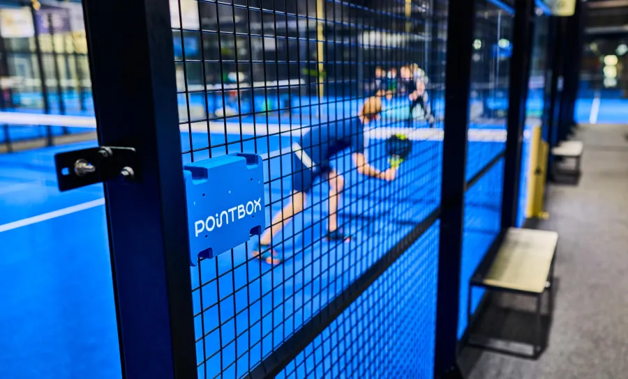 De Pointbox aan het hek van een padelbaan, terwijl er op de achtergrond gespeeld wordt